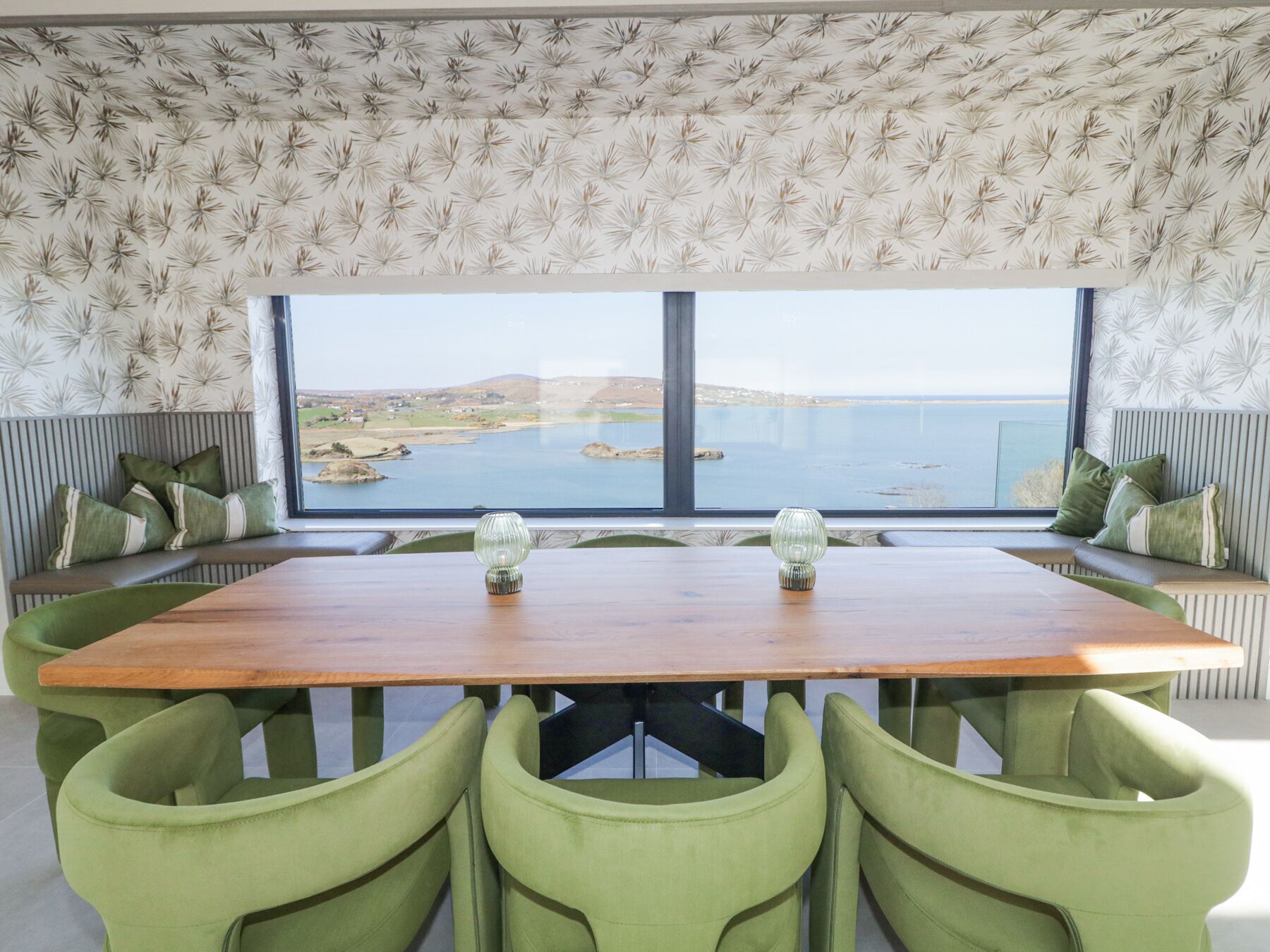 Un comedor con mesa de madera, sillas verdes y vistas a un paisaje costero a través de amplios ventanales.