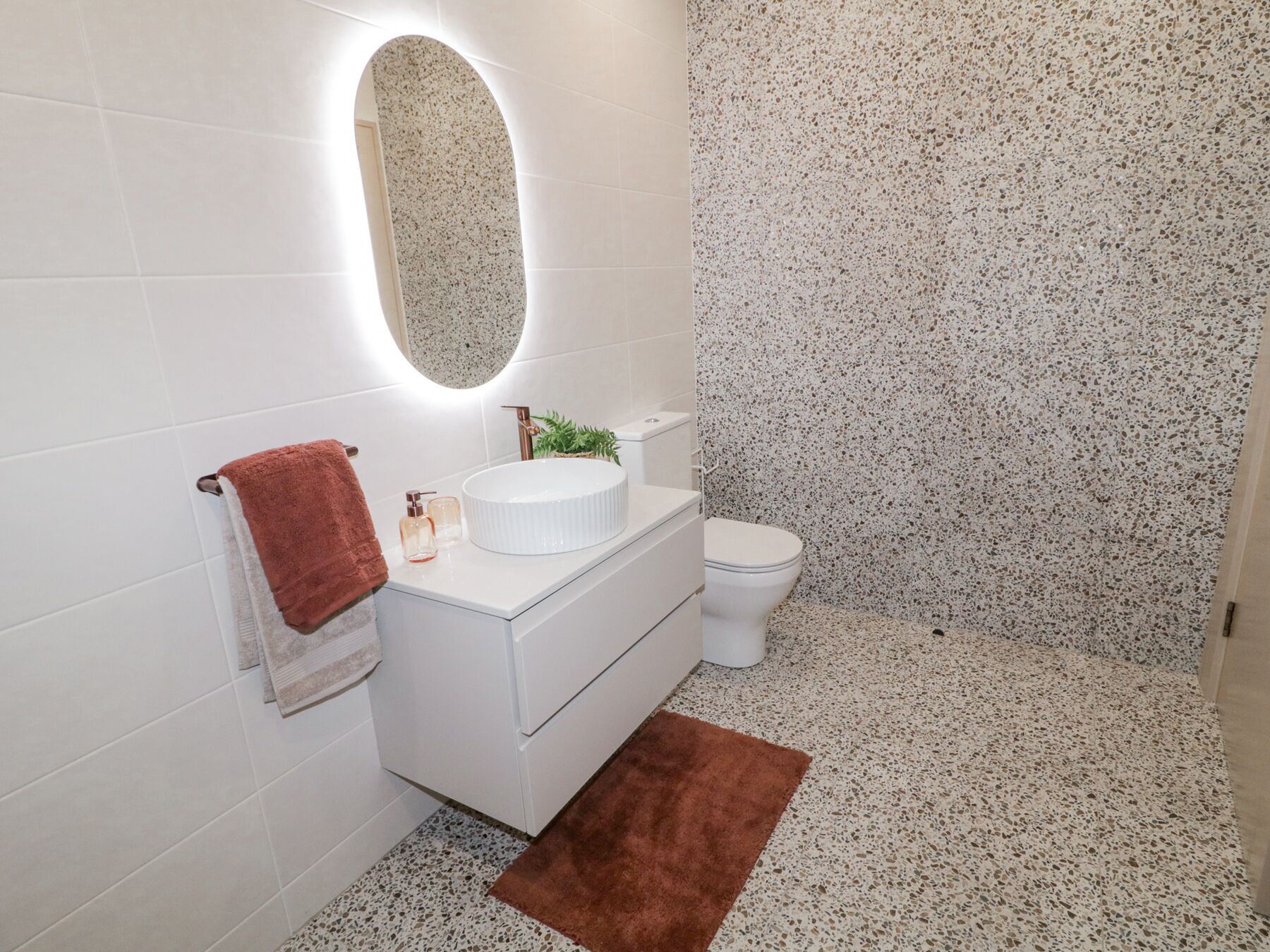 A bathroom with a white vanity, a round mirror, a toilet, and speckled flooring.