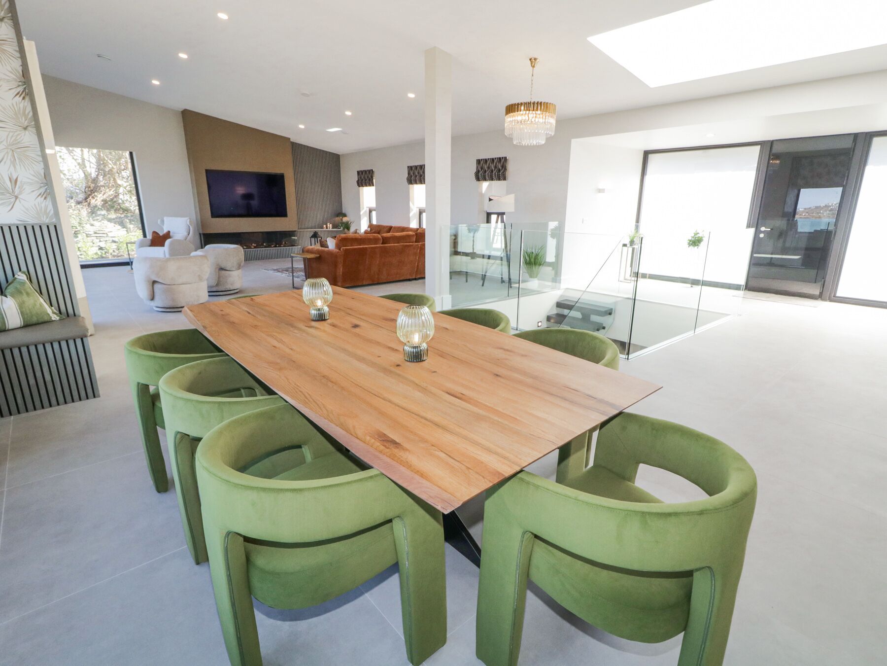 A modern dining area with a wooden table and green upholstered chairs, a television in the background, and a glass partition separating the dining area from the living space.