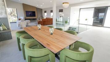 A modern dining area with a wooden table and green upholstered chairs, a television in the background, and a glass partition separating the dining area from the living space.