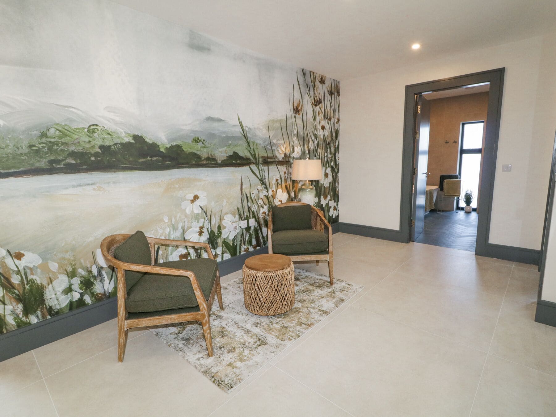 A spacious lobby with a large mural of a natural landscape, two wooden chairs, a round wooden side table, and a floor lamp.