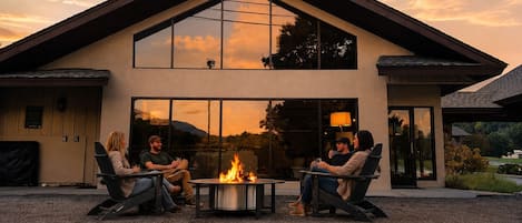 A modern house with a stone wall in front, a fire pit in the center, and people sitting around it.
