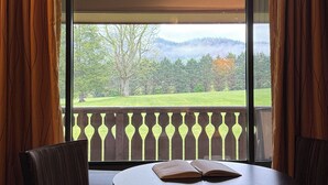 A room with a large window, a round table, two chairs, and a view of a green landscape with trees and hills.