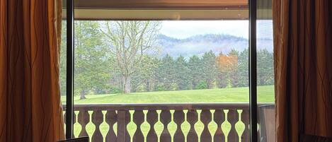 A room with a large window, a round table, two chairs, and a view of a green landscape with trees and hills.