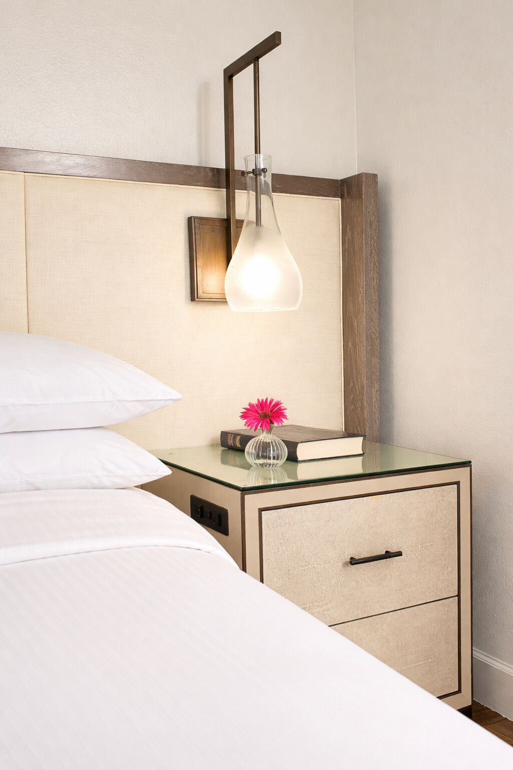 Habitación de hotel con una cama, una mesita de noche con un libro y un florero, una cómoda con cajones y una lámpara de pared.