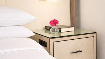 Habitación de hotel con una cama, una mesita de noche con un libro y un florero, una cómoda con cajones y una lámpara de pared.