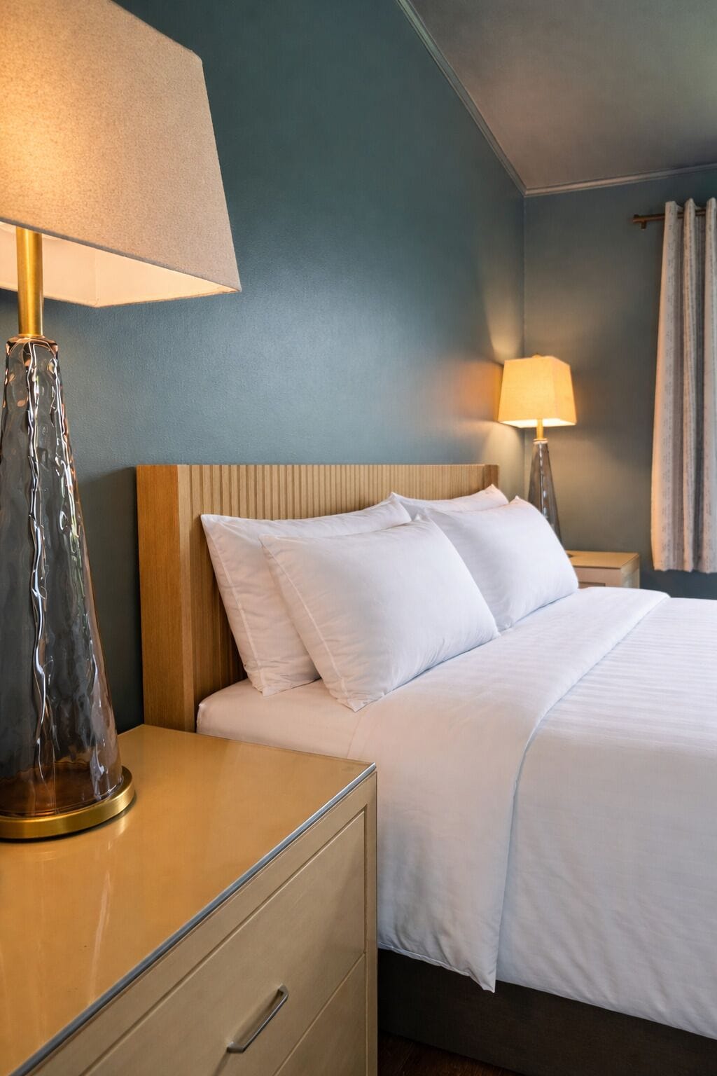 Quarto de hotel com cama, duas luminárias de cabeceira, mesa de cabeceira e uma gaveta.