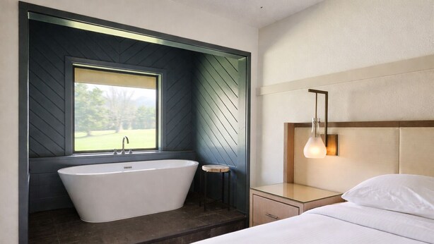 A modern bathroom with a freestanding bathtub, a window, and a bedside table with a lamp.