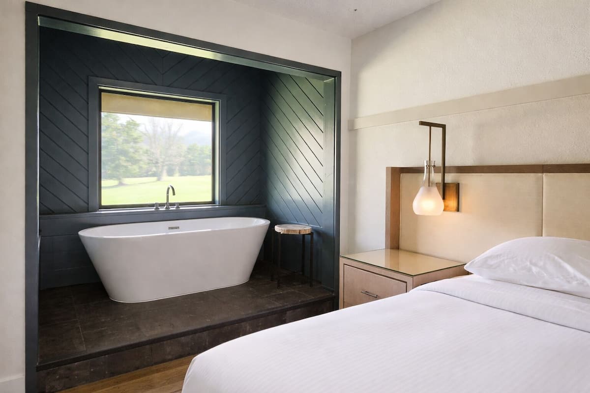 A modern bathroom with a freestanding bathtub, a window, and a bedside table with a lamp.