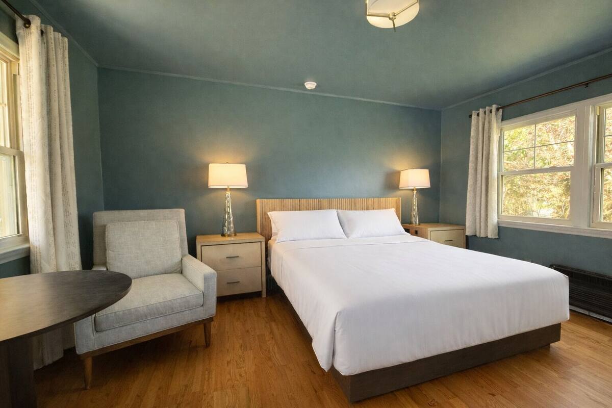 A hotel room with a bed, a chair, a table, two bedside lamps, and a window.