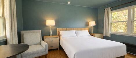 A hotel room with a bed, a chair, a table, two bedside lamps, and a window.