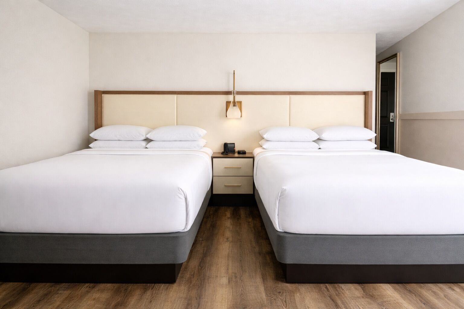 Habitación de hotel con dos camas, una mesita de noche con lámpara y suelo de madera.