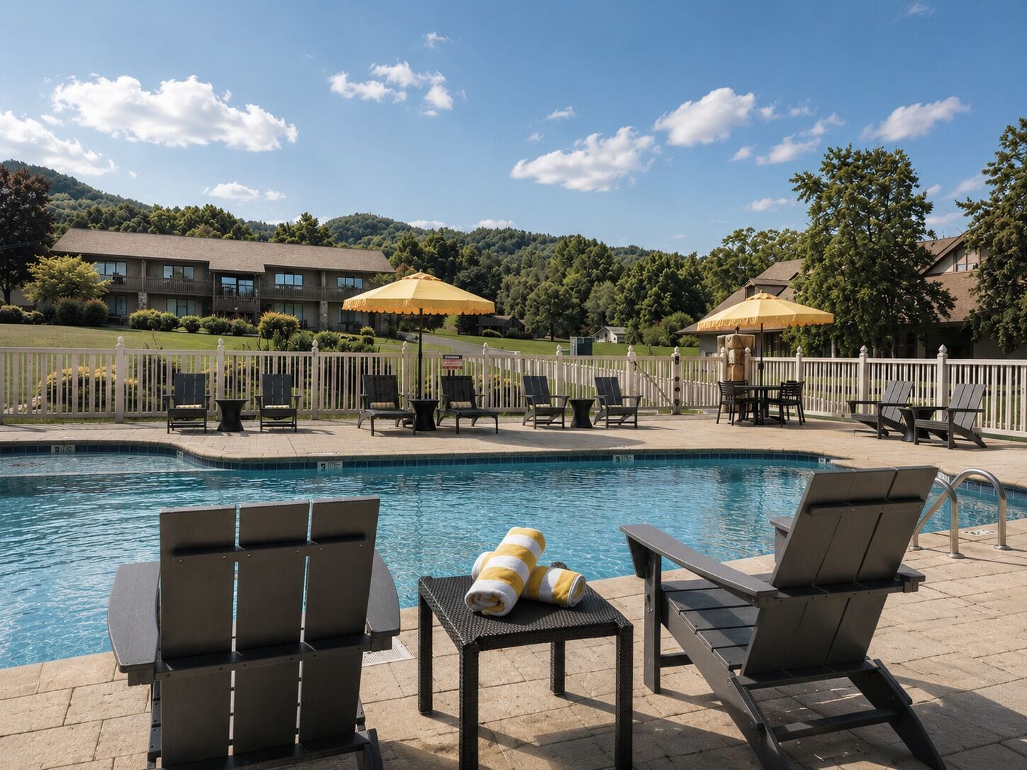 A pool area with lounge chairs and umbrellas, surrounded by a fence and buildings in the background.