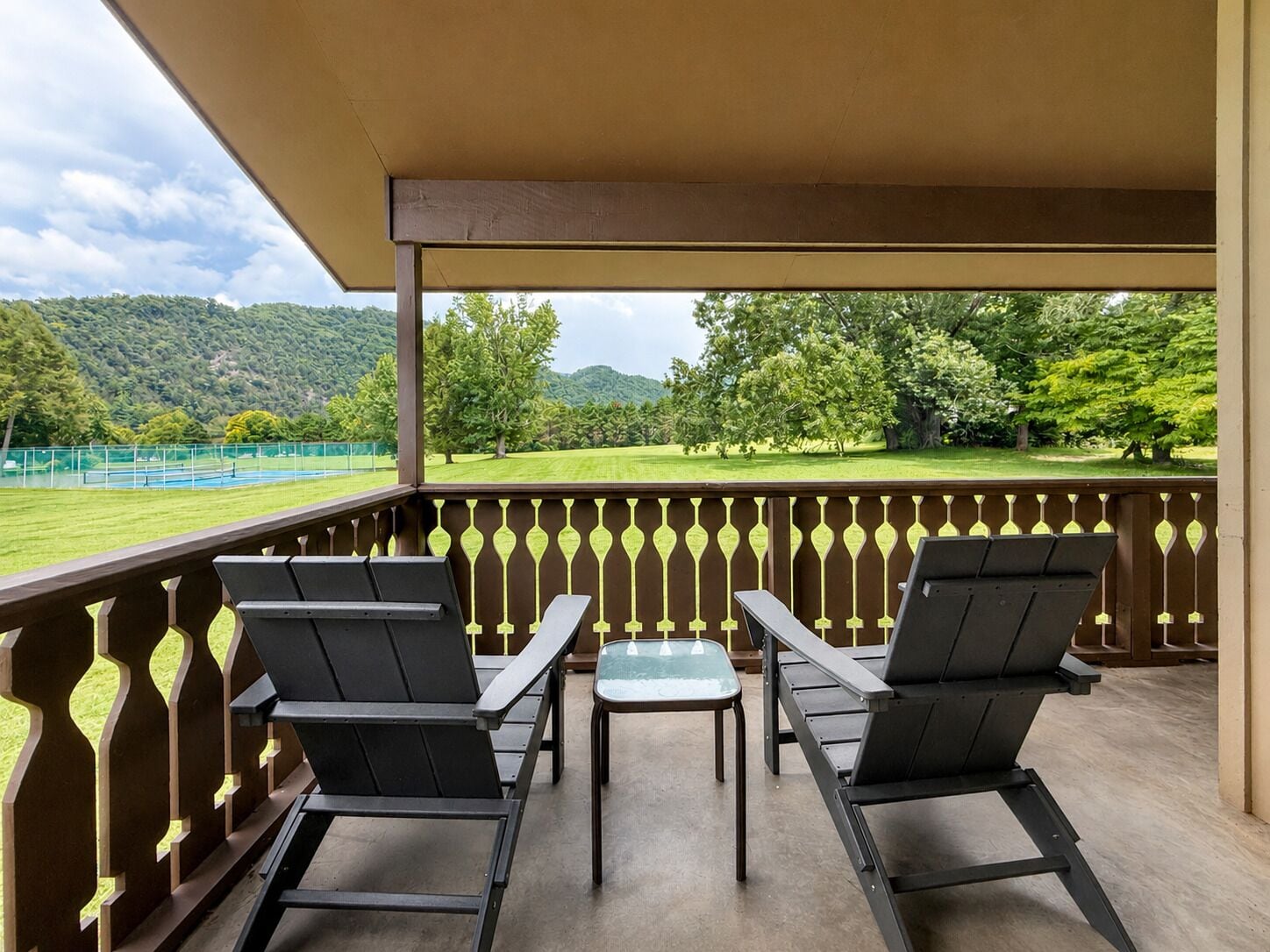 A balcony with two chairs and a small table, overlooking a green landscape with trees and a tennis court.