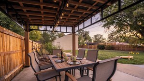 An outdoor patio with a wooden table set for four, surrounded by chairs, under a pergola with hanging lights. - Mile High Retreat - Hot tub/Huge Backyard/Pool Table/Ping Pong/BoardGames/Arcade (Denver)