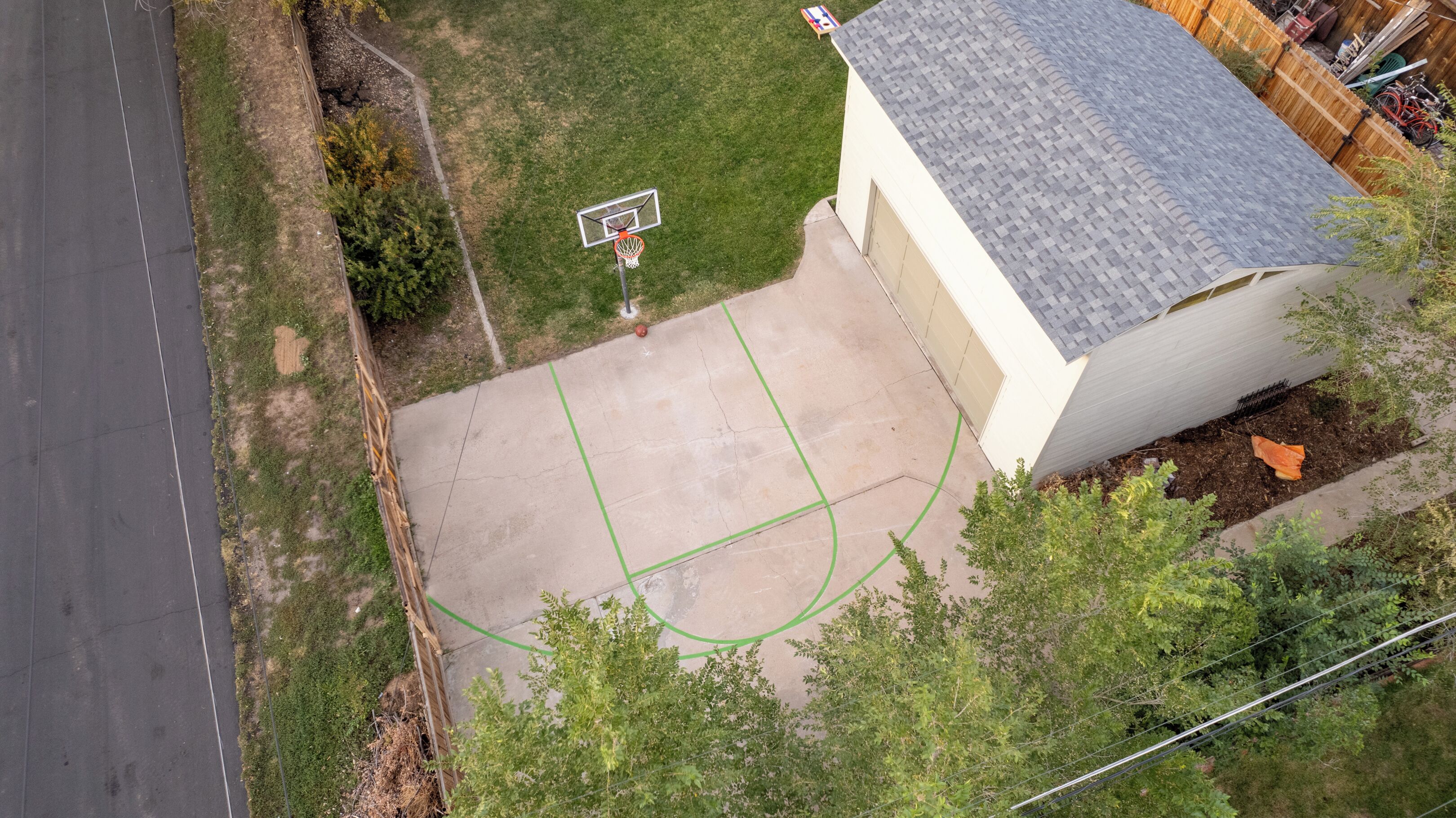 Vue aérienne d’un quartier résidentiel avec un terrain de basketball, une maison avec un toit gris et une clôture en bois.