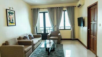 A living room with a sofa, armchair, and coffee table. There are two windows with curtains, an air conditioning unit, and a television mounted on the wall.