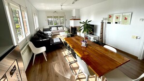 A modern living room with a dining area, featuring a wooden dining table, white chairs, a black sofa, and a ceiling fan.