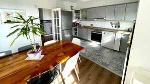 A modern kitchen with white cabinets, a built-in oven, and a dishwasher. There is a dining table with chairs and a potted plant.