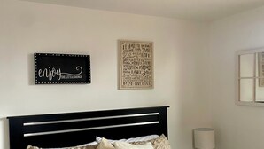 Een net opgemaakt bed met een gewatteerde spreideken, een nachtkastje met een lamp, en twee motiverende kunstwerken aan de muur.