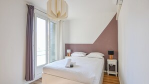 A modern bedroom with a large bed, white curtains, a ceiling light, and a nightstand with a lamp.