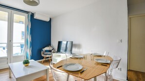 A dining area with a wooden table, clear chairs, and a white bench. There is a small plant on the bench, and a TV is mounted on the wall.