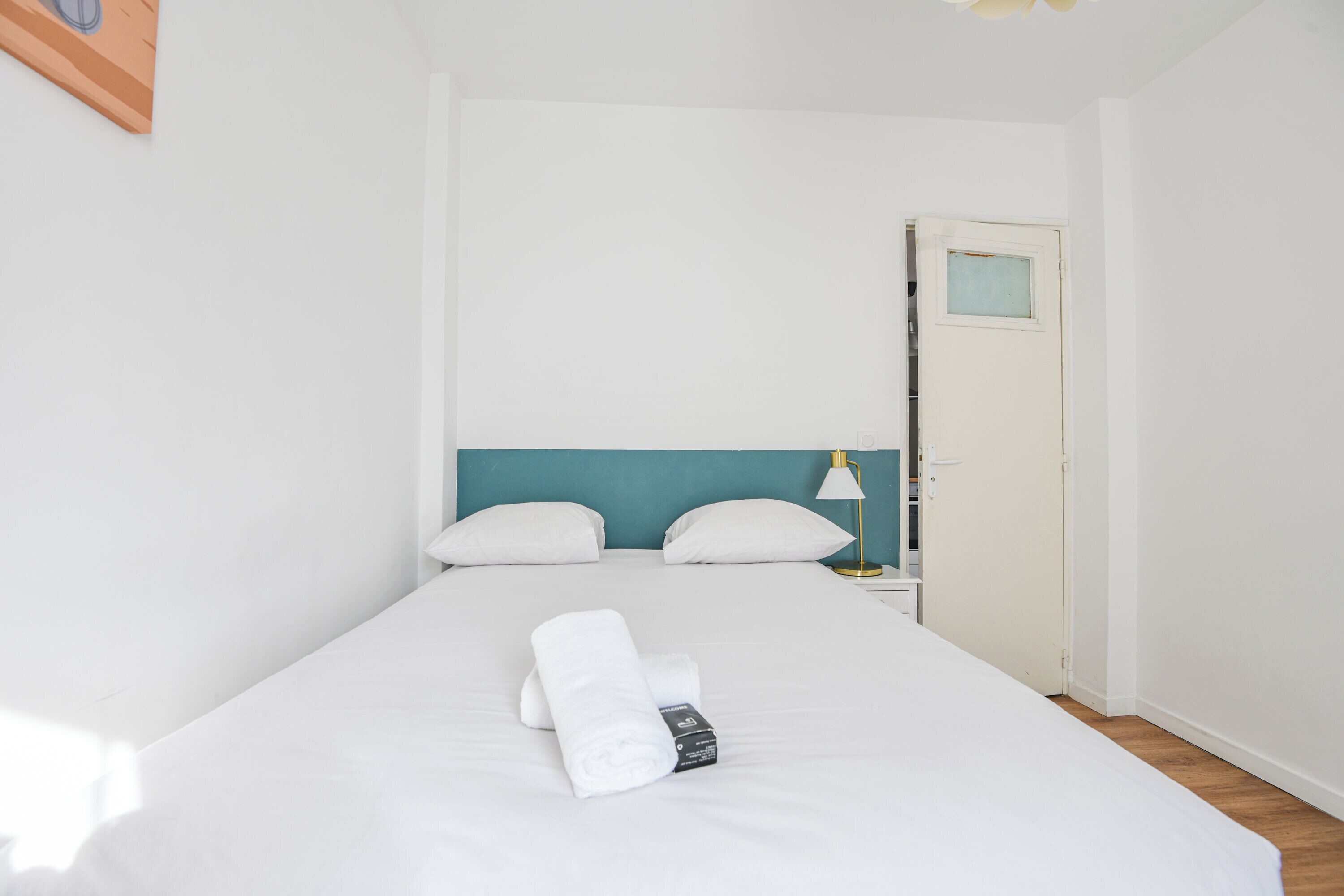 A neatly made bed with white linens and a rolled towel, a bedside lamp, and a door with a small window.