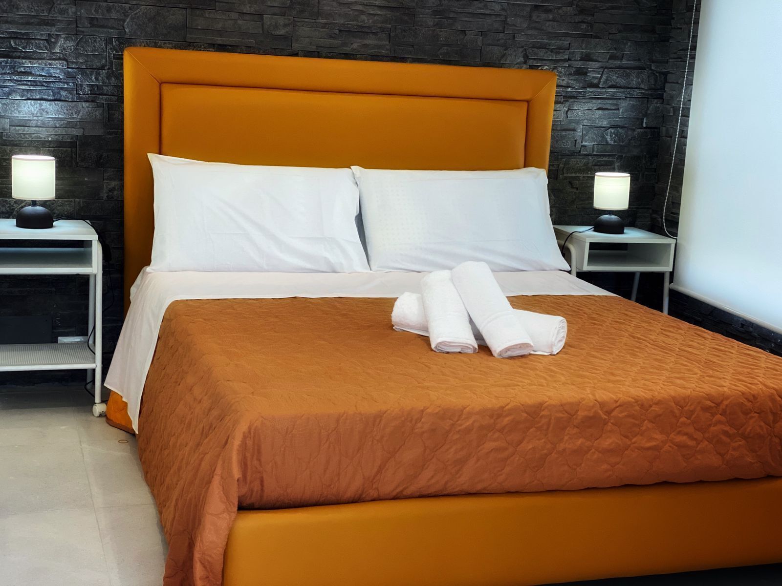Un lit bien fait, avec une tête de lit marron, des oreillers blancs et une serviette pliée posée sur le lit. Deux tables de chevet, chacune avec une lampe, se trouvent de part et d’autre du lit.