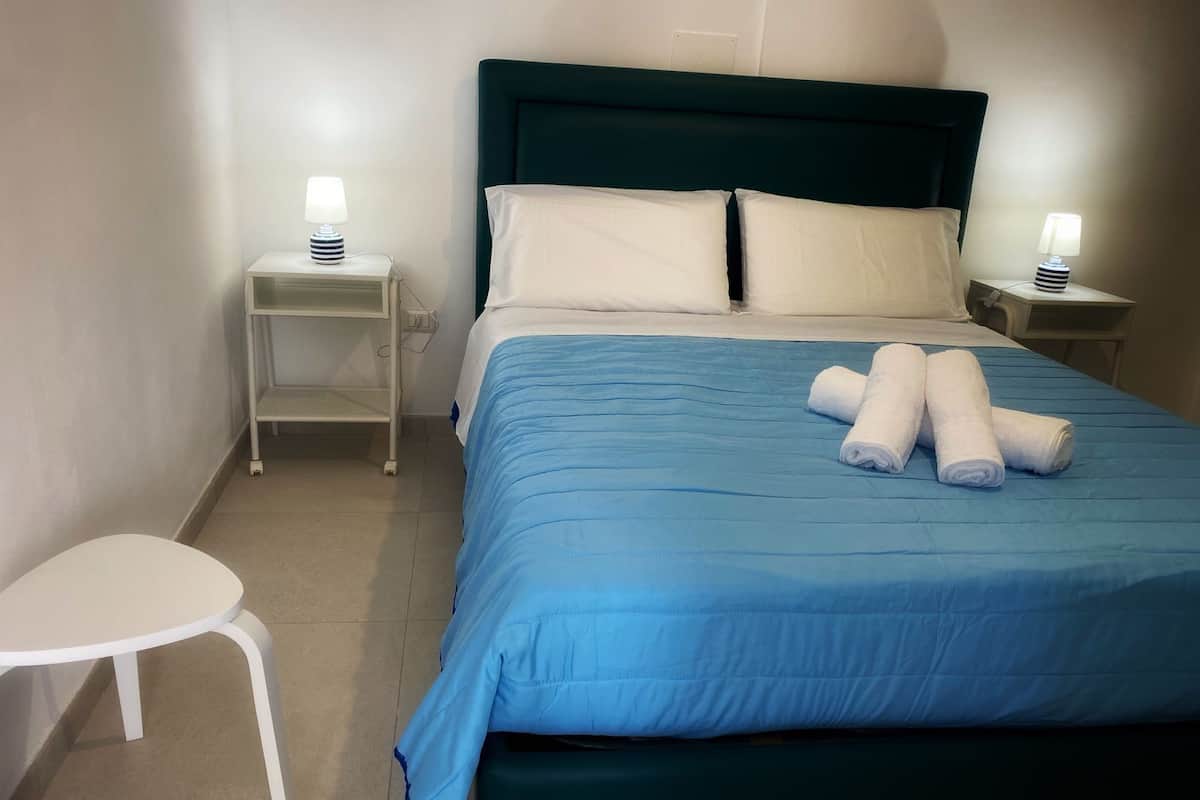 En pænt redt seng med blåt sengetøj og hvide puder, to sengeborde med lamper, og en hvid stol.