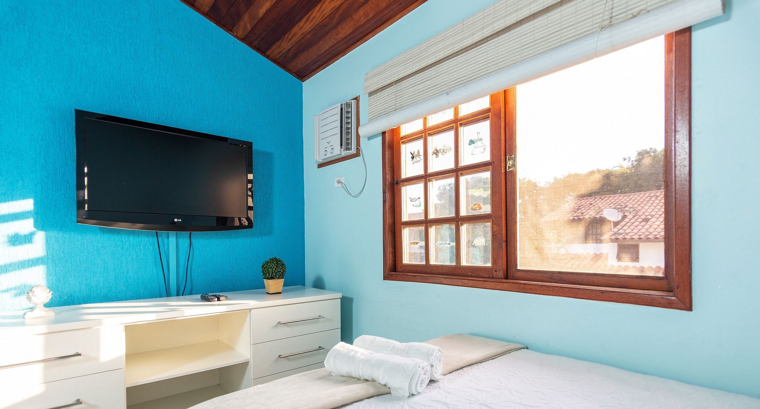 Una stanza con una parete blu, un soffitto in legno, un letto con biancheria bianca, una scrivania bianca con cassetti, un televisore a schermo piatto montato al muro e una finestra con vista sugli edifici.