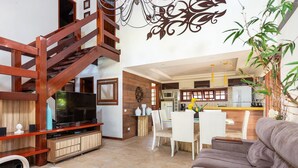 Amplia sala de estar con escalera de madera, televisor de pantalla plana, mesa de comedor con sillas y cocina al fondo.