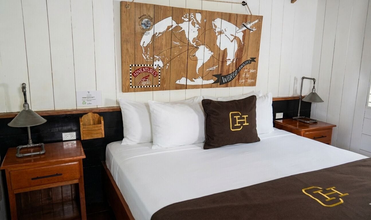 Een hotelkamer met een bed, twee nachtkastjes, een wereldkaart als kunstwerk, een kussen met een logo en een bordje aan de muur.