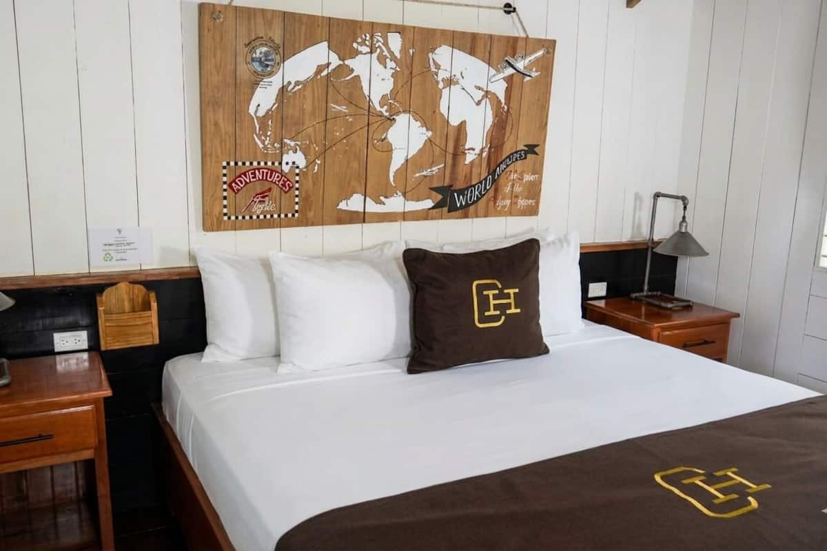 Una habitación de hotel con cama, dos mesitas de noche, una obra de arte con un mapa del mundo, un almohadón con un logotipo y una señal en la pared.
