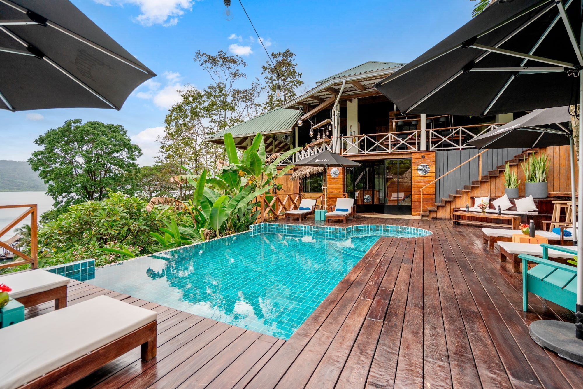 A wooden deck with a swimming pool, lounge chairs, and a covered seating area with a view of the water.