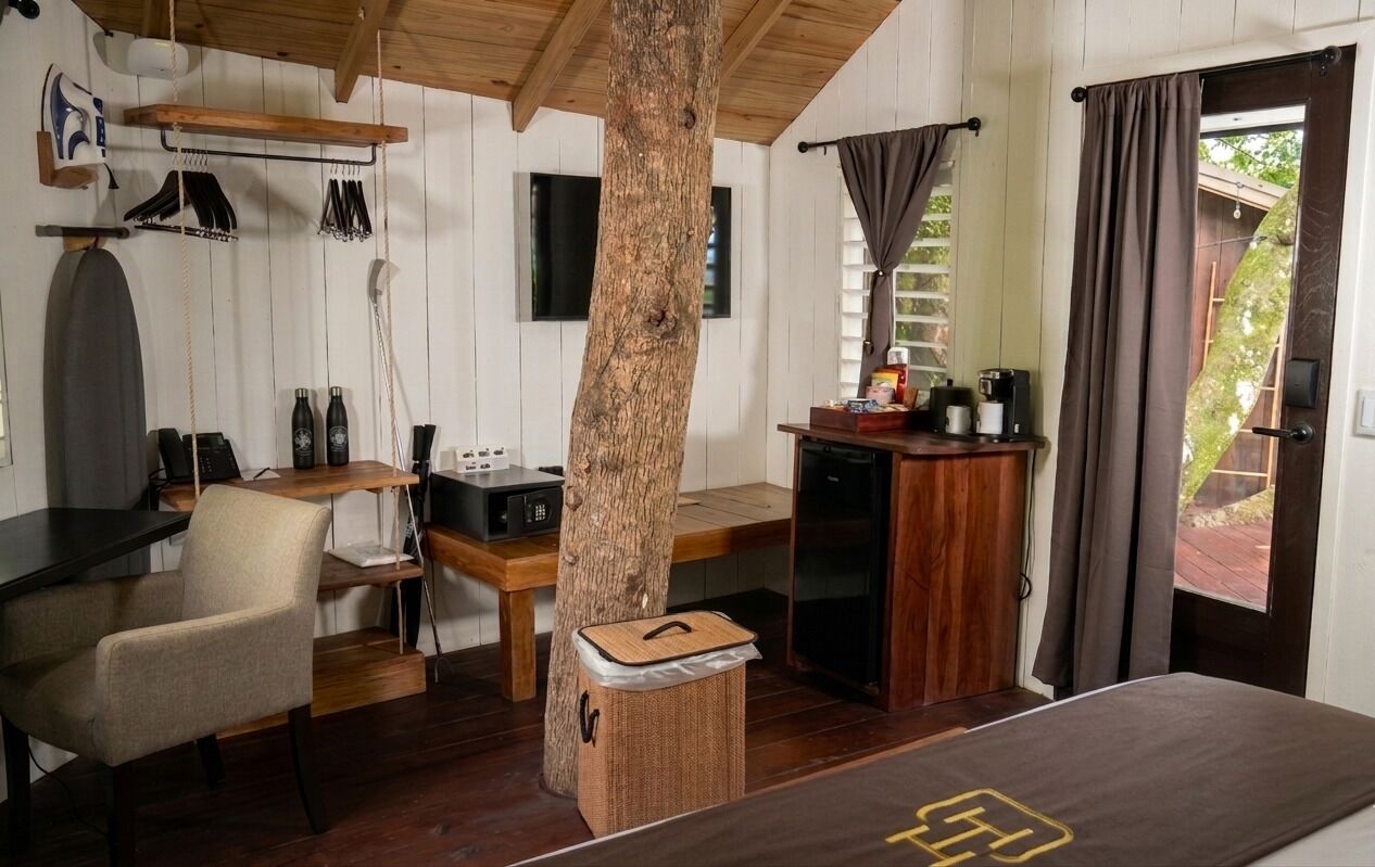 A compact room with a wooden desk, a chair, a small kitchenette, a bed, and a large tree trunk centerpiece.