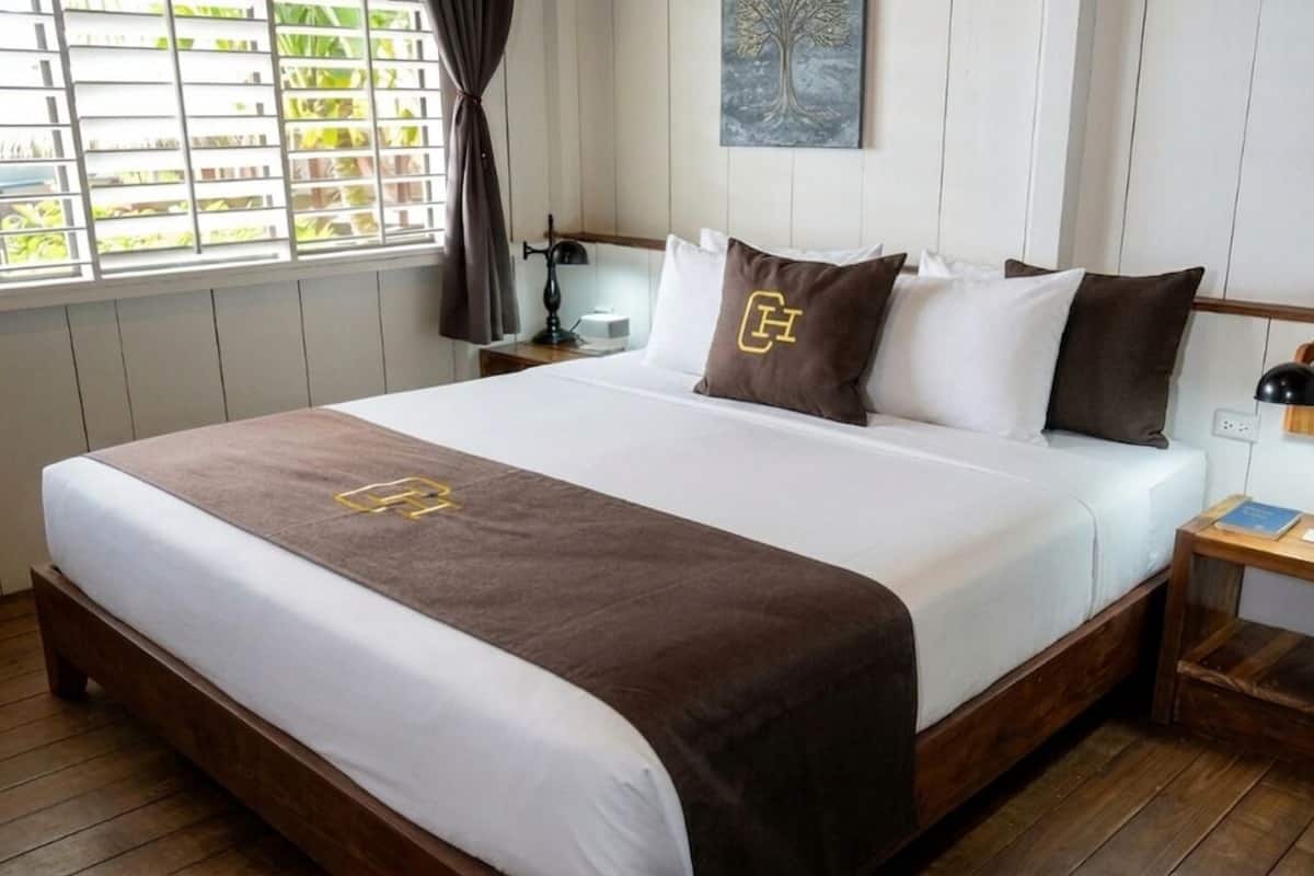 Een net opgemaakt bed met een bruine sprei en een logo, een nachtkastje met een lamp, en een raam met zonwering.