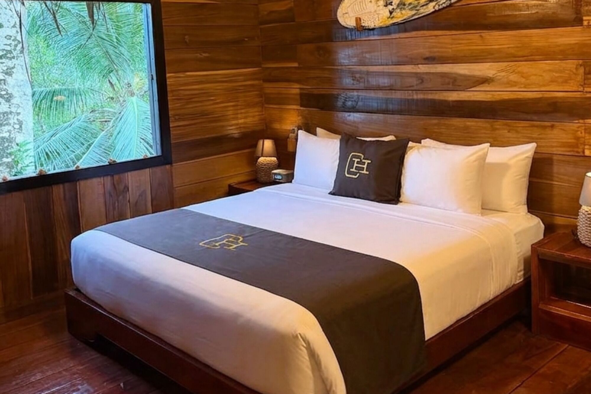 Een houten kamer met een bed, een nachtkastje, een lamp en een groot raam met uitzicht op palmbomen.