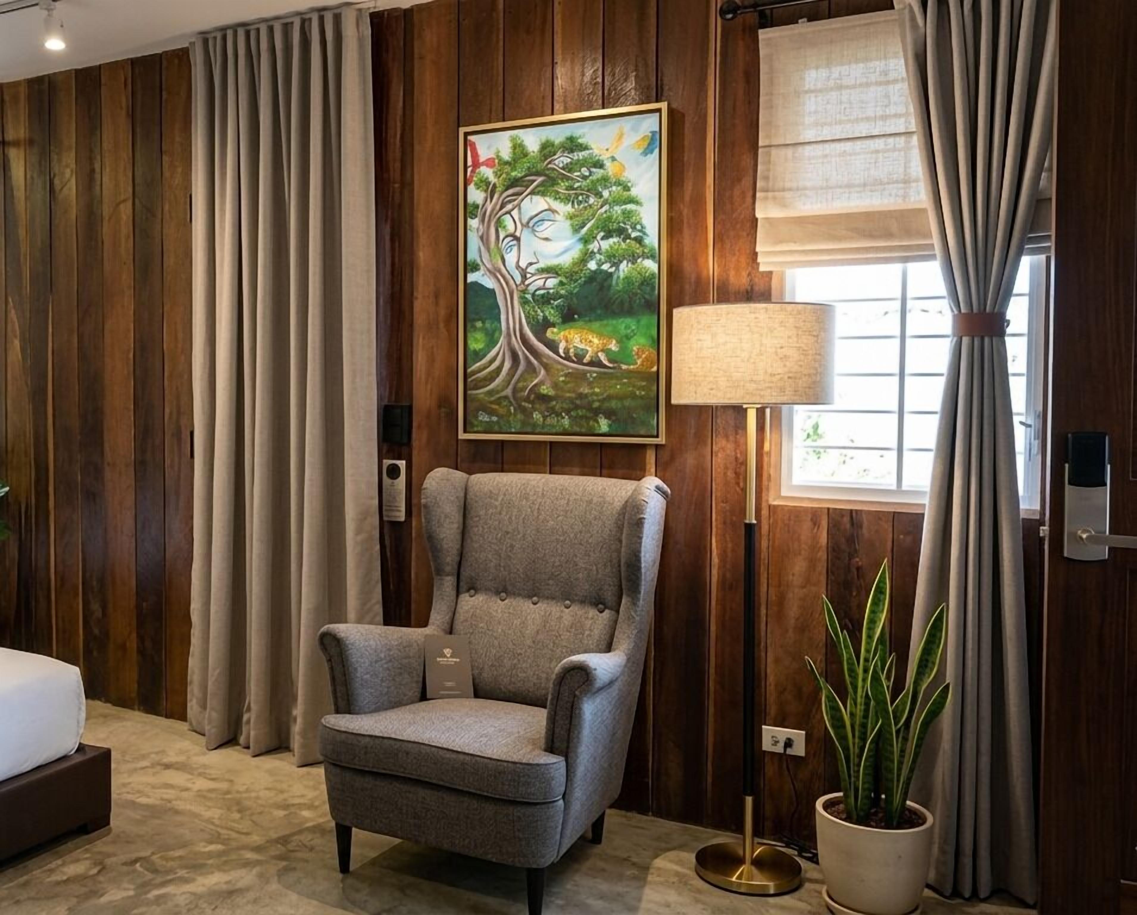 A room with a grey armchair, a painting of a tree and leopard, a lamp, and a potted plant.