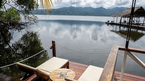 A wooden deck with a table and two lounge chairs overlooking a calm lake with mountains in the background. - Central Hotel Bocas del Toro (Bocas del Toro)