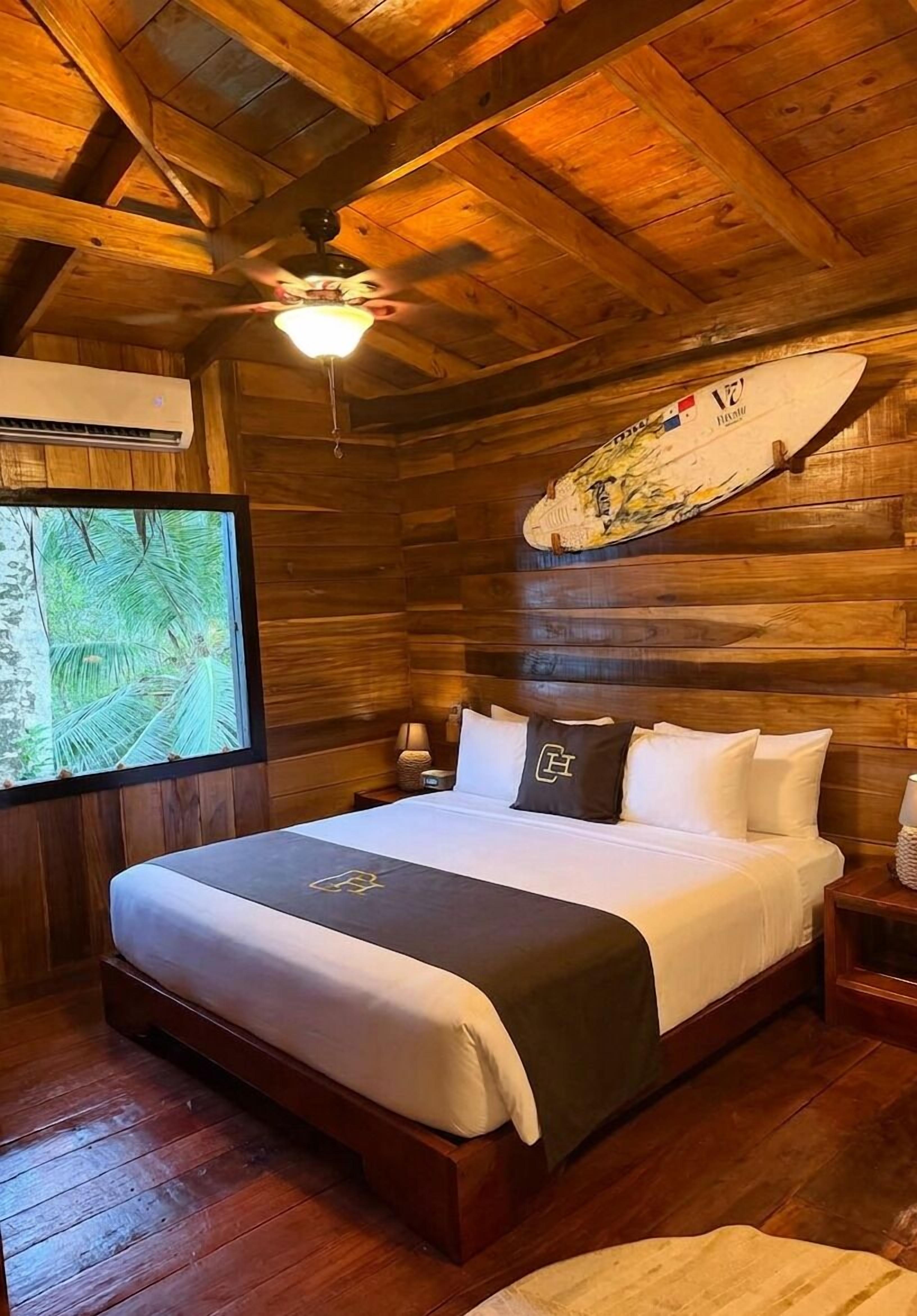 Ein holzgetäfeltes Zimmer im Blockhausstil mit einem Bett, einem Deckenventilator, einem an der Wand befestigten Surfbrett und einem großen Fenster mit Blick auf eine tropische Landschaft.