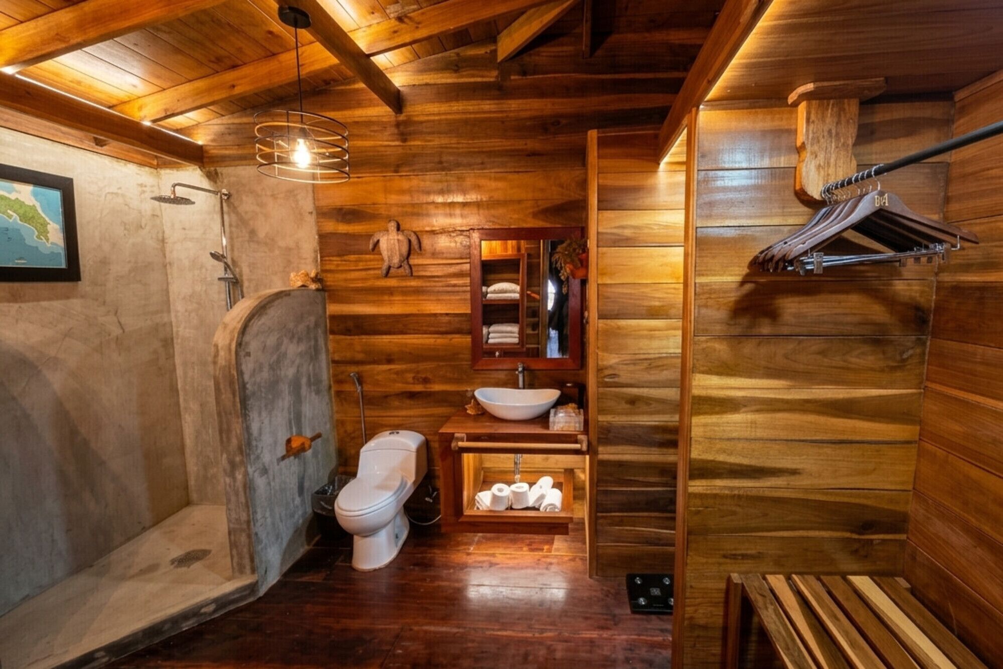 Une salle de bain rustique, avec des murs en bois, comprenant un banc en bois, des toilettes, un lavabo, un miroir et un porte-serviettes.