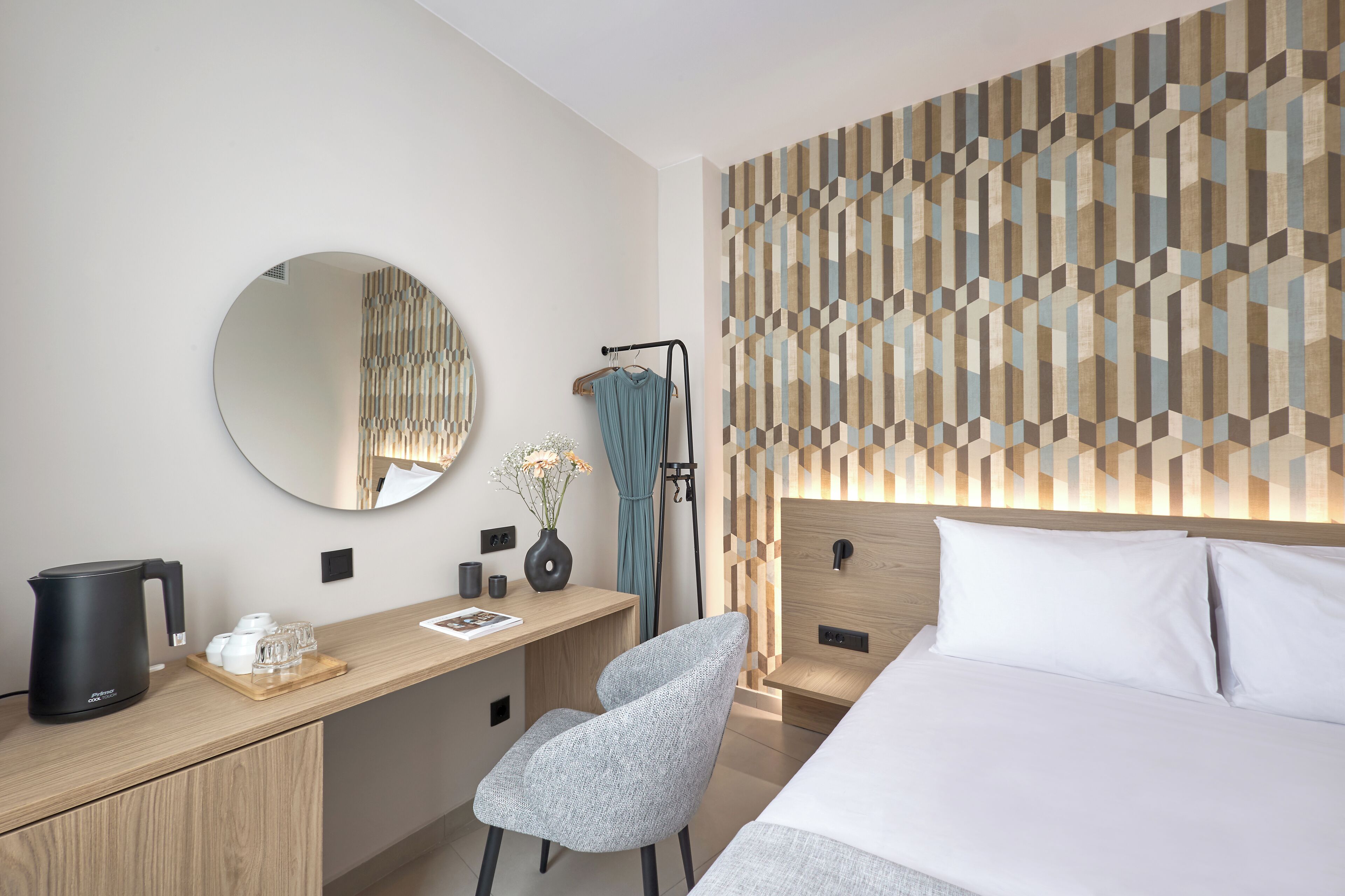 A modern hotel room with a wooden desk, a round mirror, a bed with white linens, and a geometric patterned headboard.