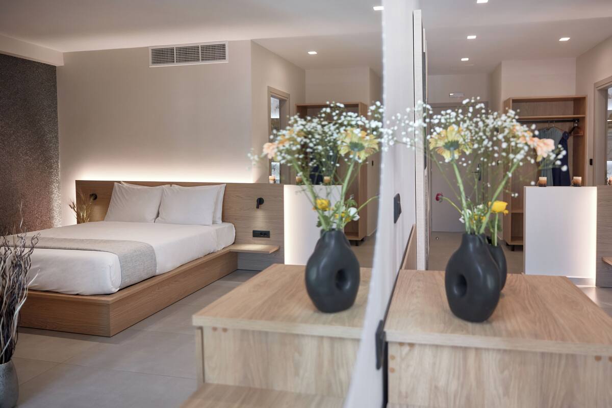 Quarto de hotel moderno com uma cama grande, mesas de cabeceira, um espelho e dois vasos pretos com flores.