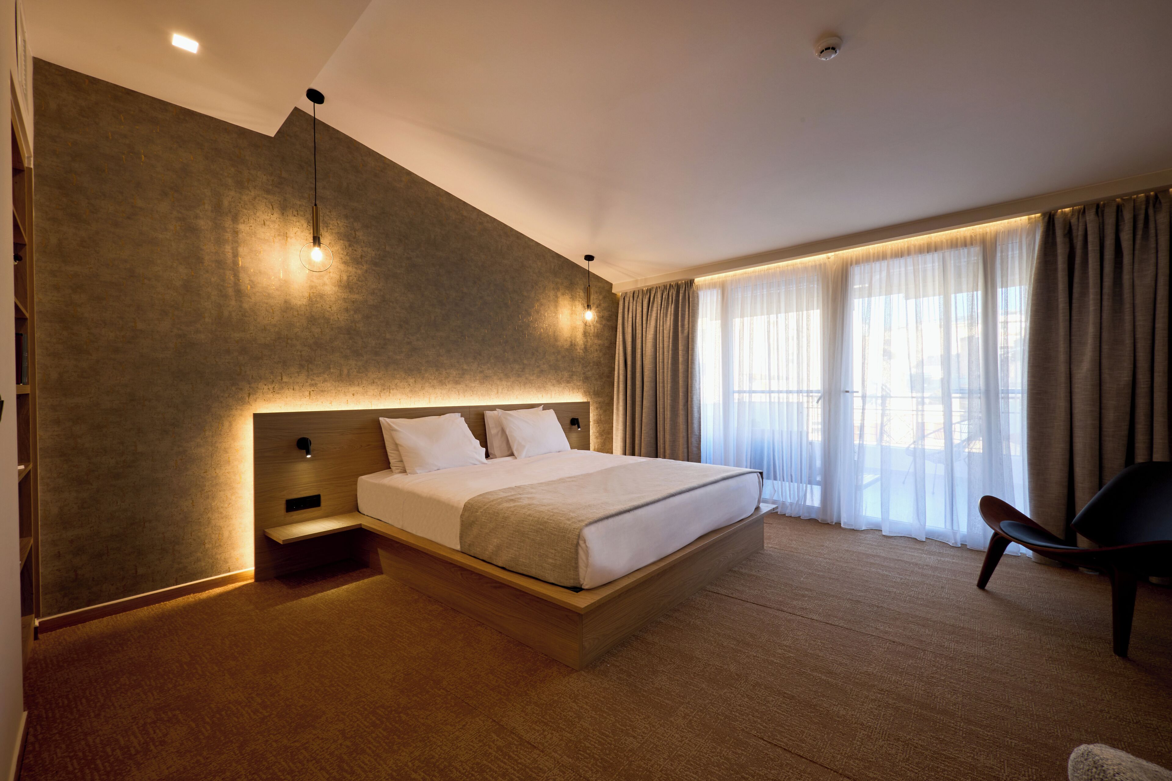 A modern hotel room with a large bed, a wooden headboard, a bedside table, and a chair.