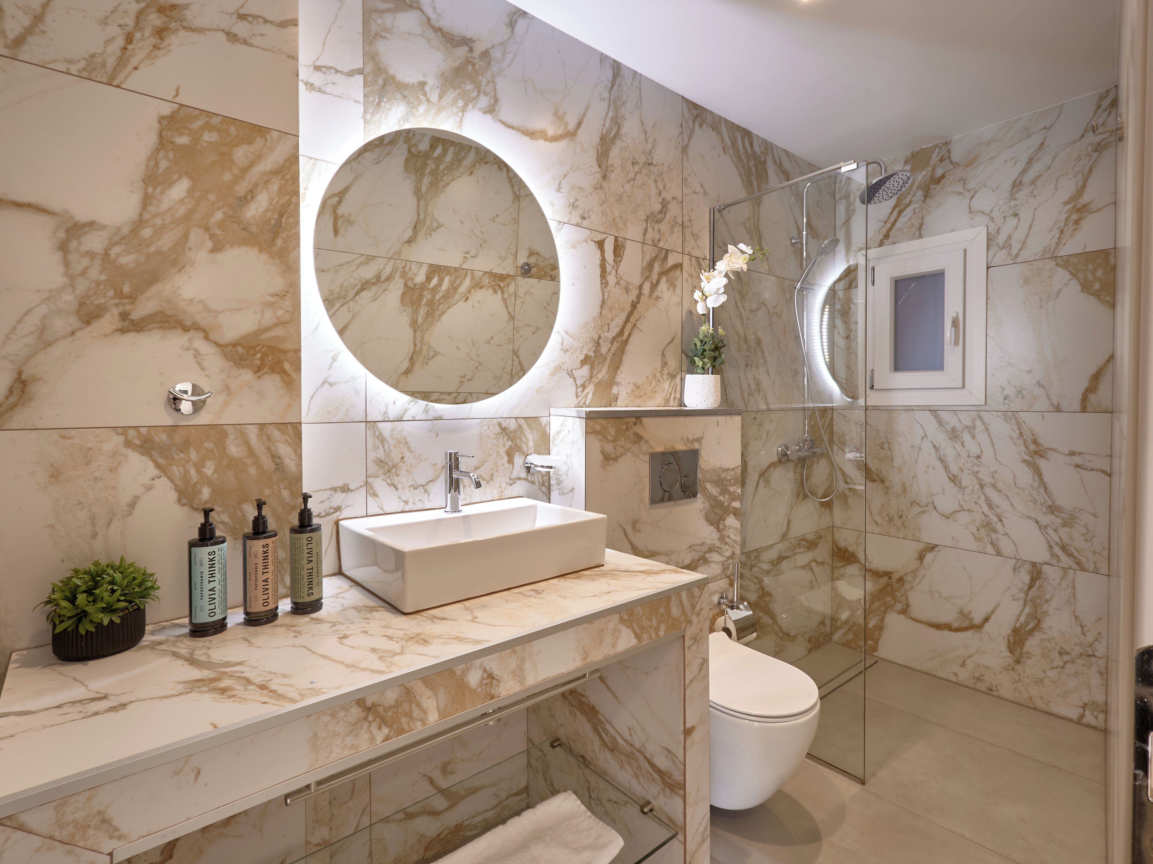 A modern bathroom with a marble vanity, a large round mirror, a glass shower enclosure, and a toilet.