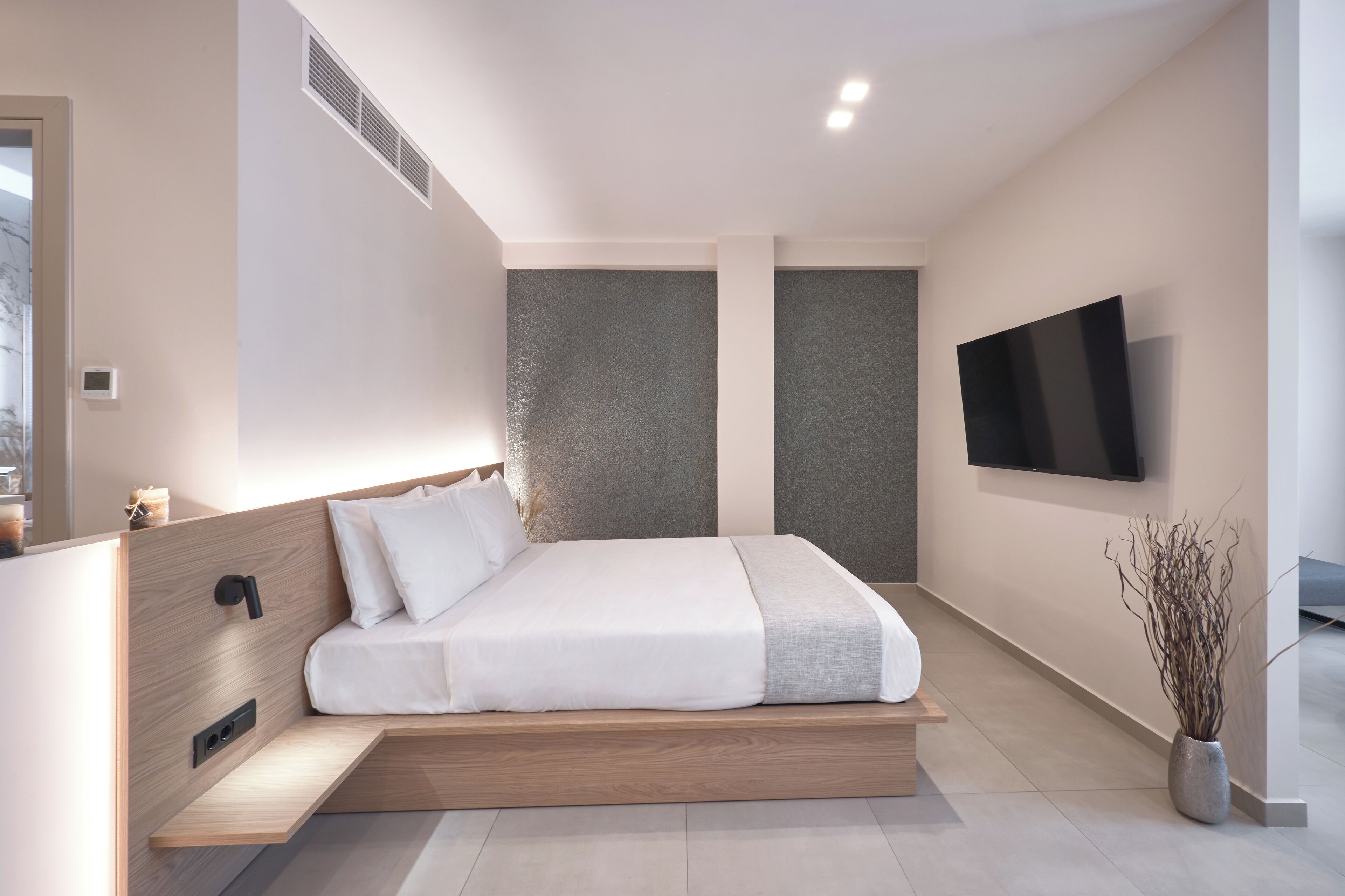 A modern bedroom with a minimalist design, featuring a bed with a wooden headboard, a flat-screen TV mounted on the wall, and a small potted plant.