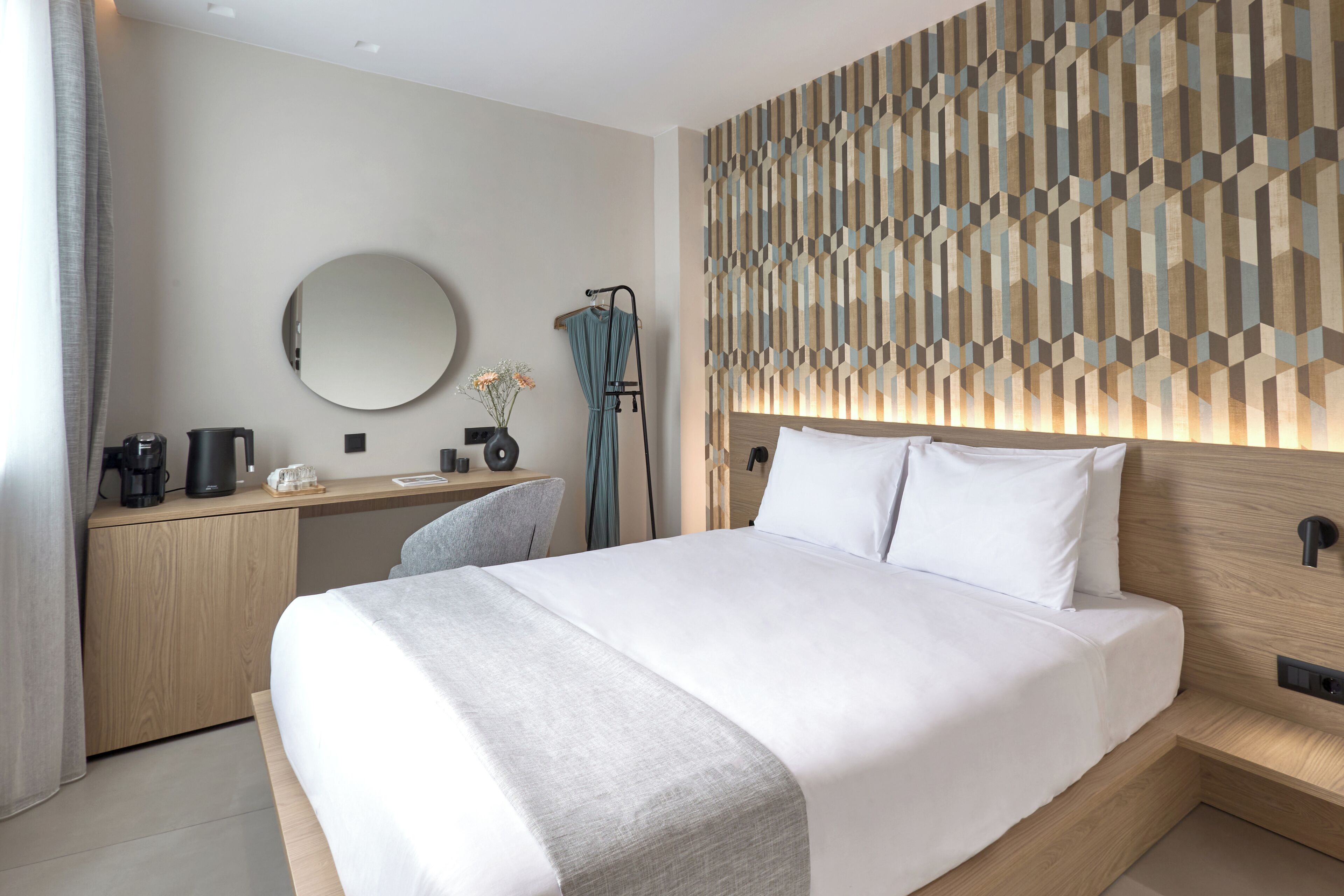 A modern hotel room with a large bed, a wooden desk, a round mirror, and a geometric-patterned wall.