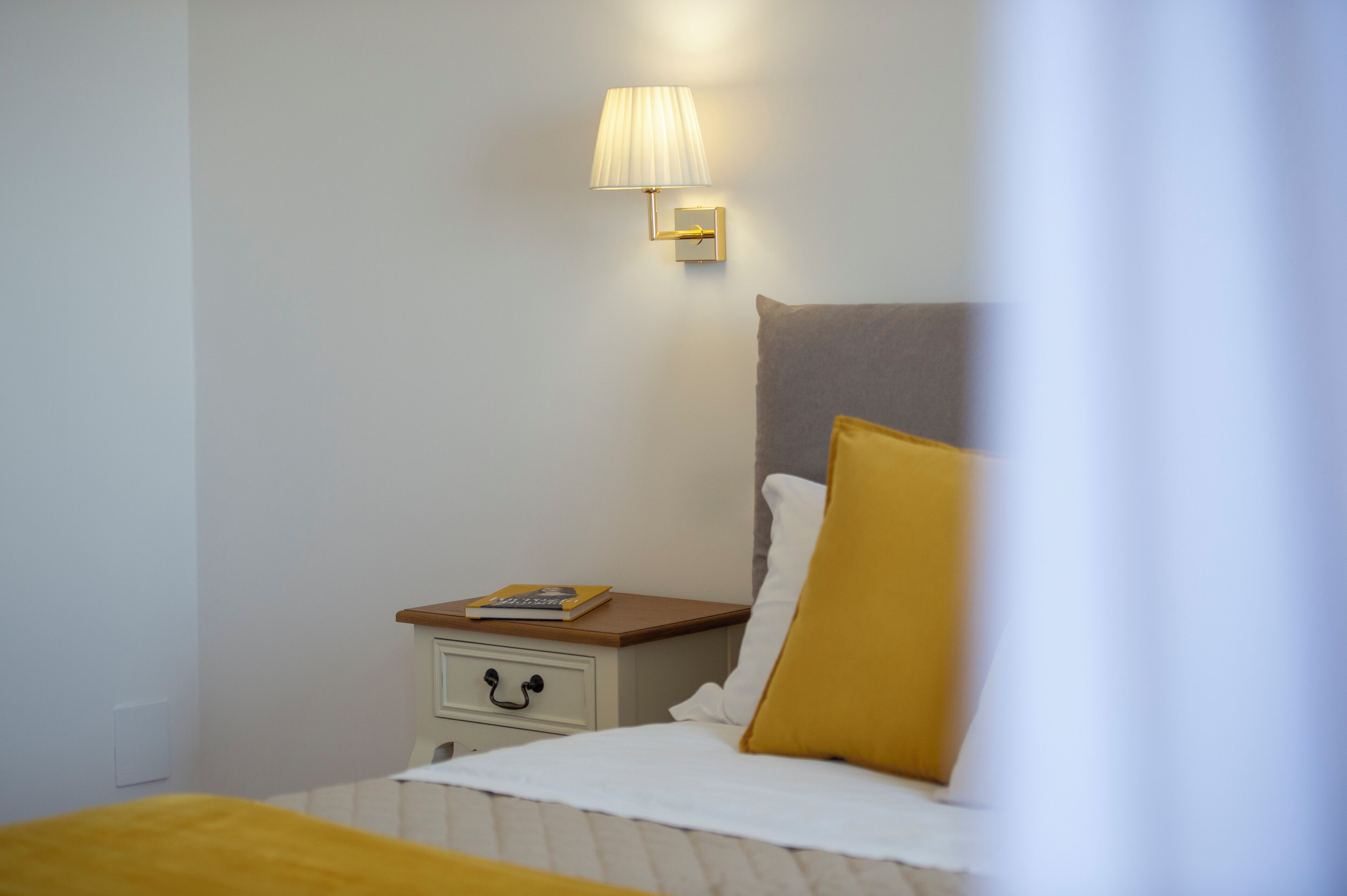 A neatly made bed with a white and yellow pillow, a bedside table with a book, and a wall-mounted lamp.