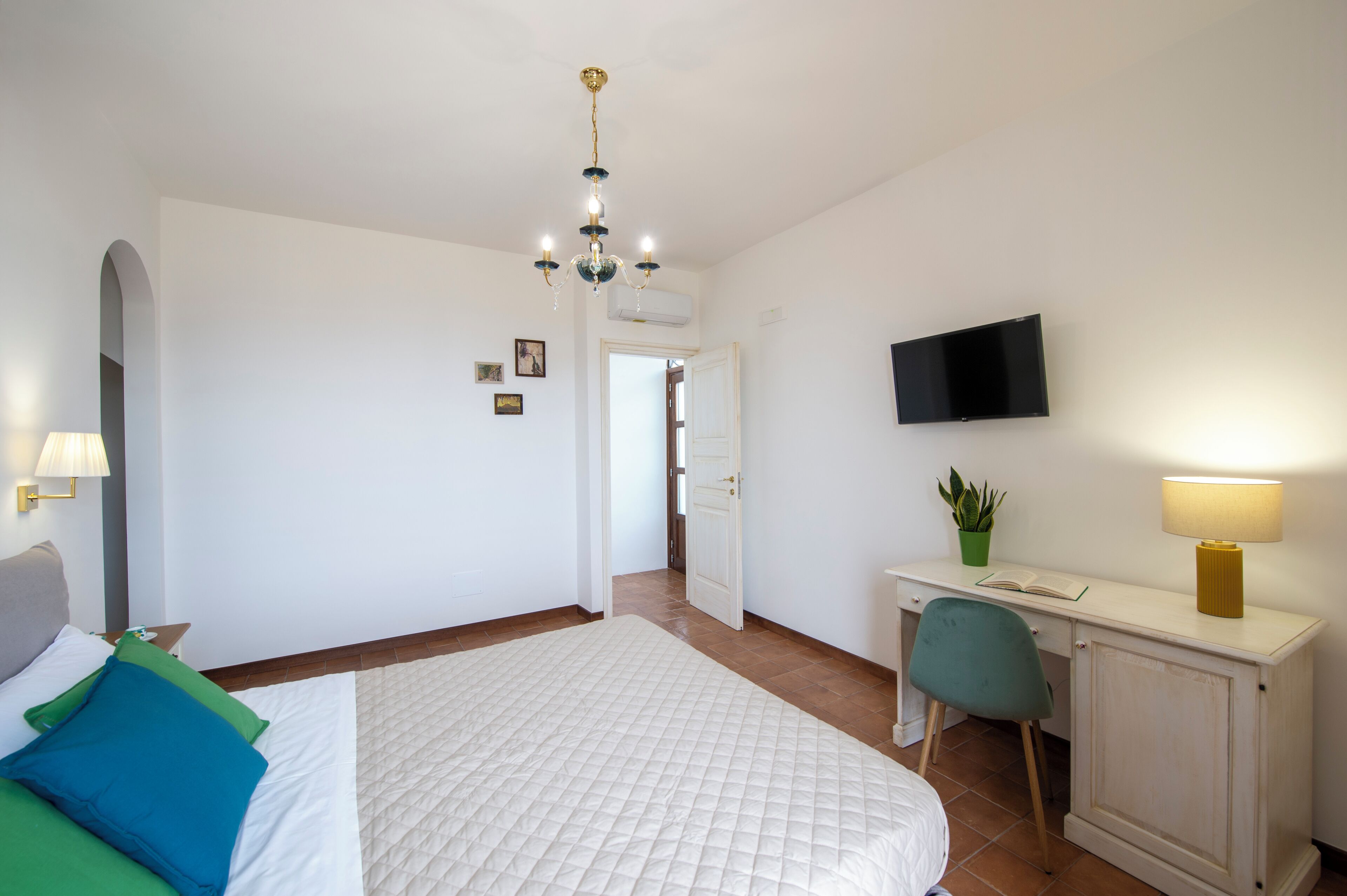 Hotellihuone, jossa on sänky, työpöytä, tuoli, televisio ja kattokruunu.