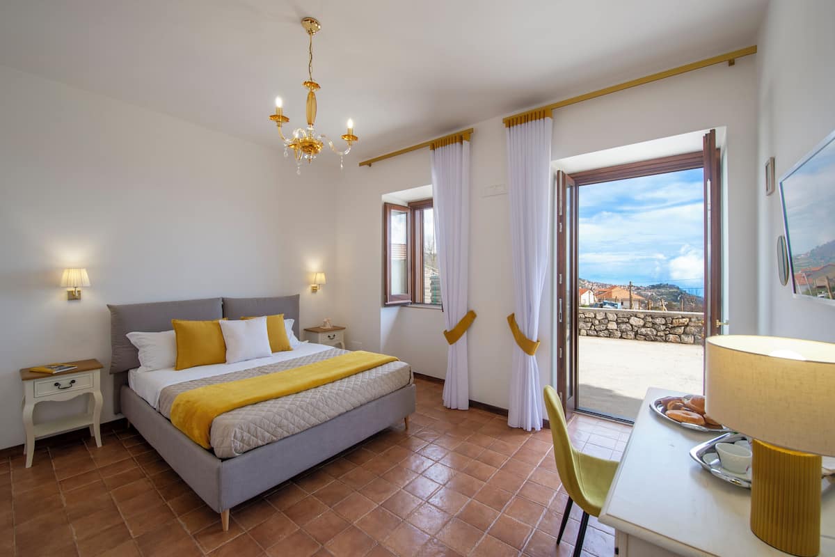 A bedroom with a bed, bedside tables, a chandelier, curtains, a balcony with a view, and a dining table with a lamp.